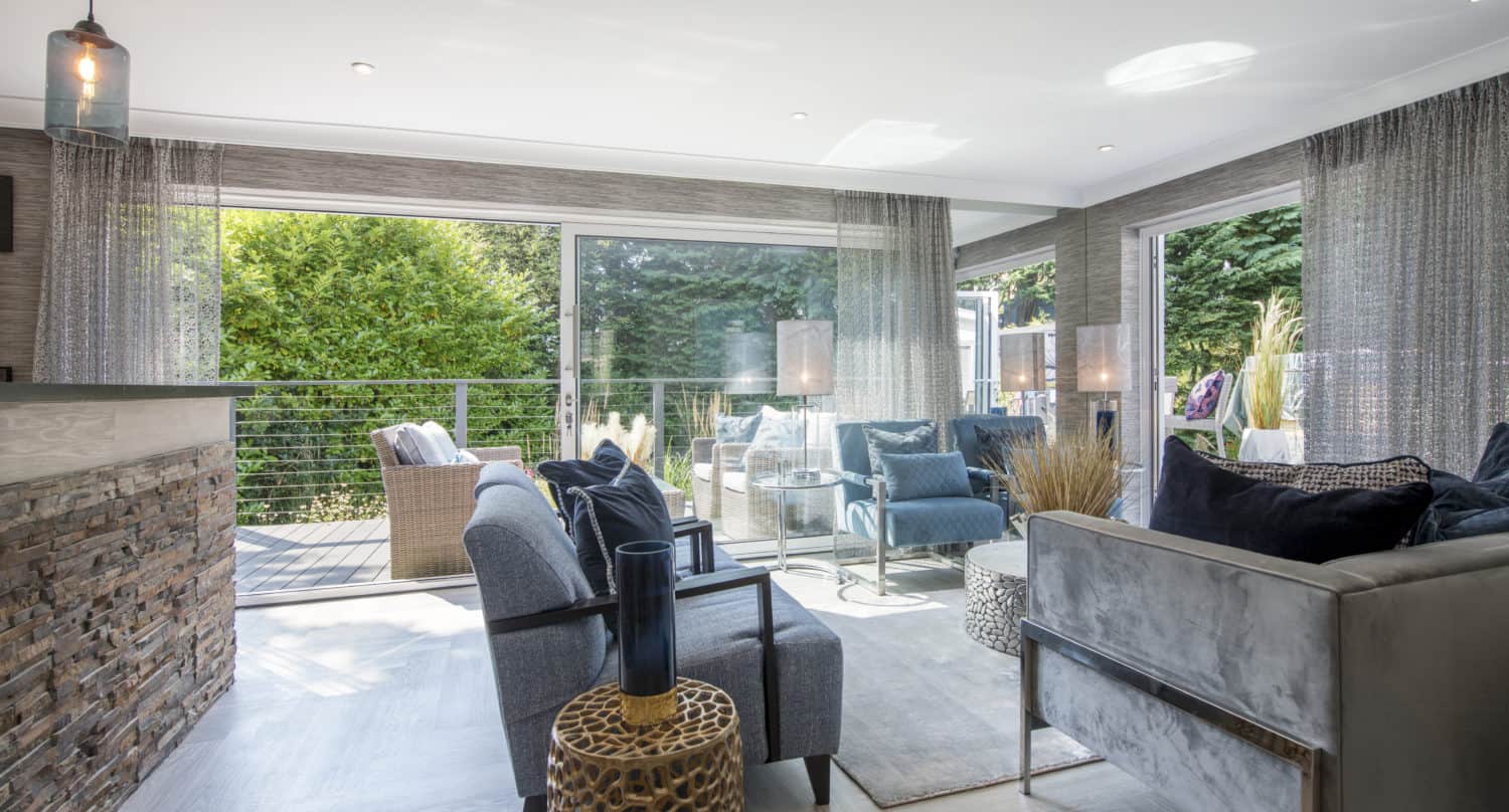 Modern living room with large windows and sliding doors opening to a green garden. The space is furnished with blue and gray sofas, accent tables, and decorative pillows, creating a bright and inviting atmosphere.