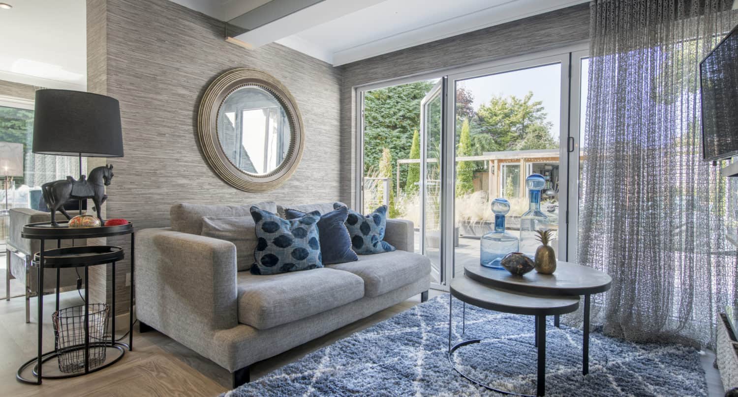 A modern living room with a gray sofa, blue patterned pillows, a round mirror, a round coffee table, and large sliding doors opening to a garden. Light filters through sheer curtains onto a blue rug.