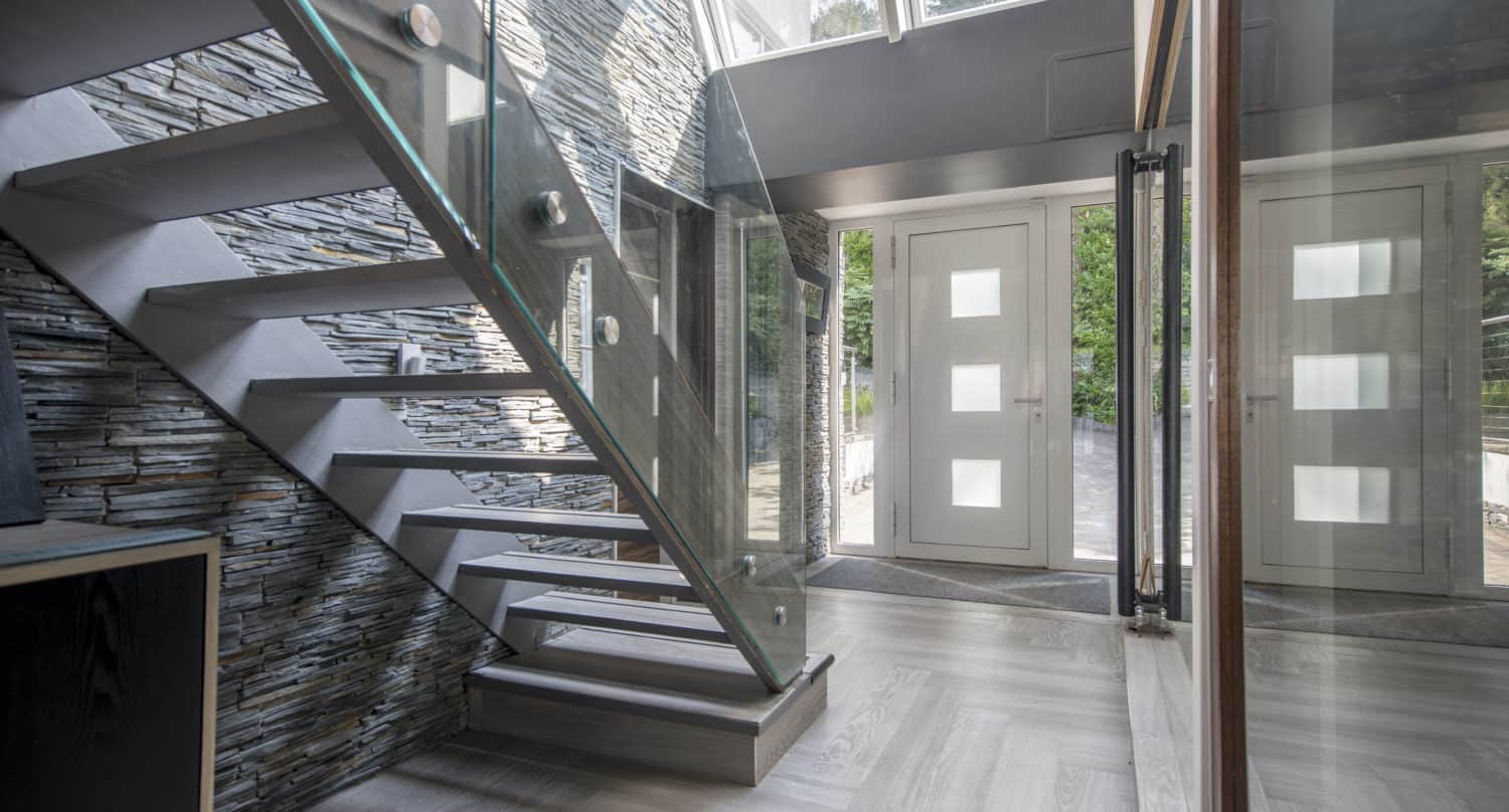Modern entryway with floating wooden stairs and glass railing, stone accent wall, bespoke glass solutions for the ceiling panels, large white double front doors with square windows, and abundant natural light.