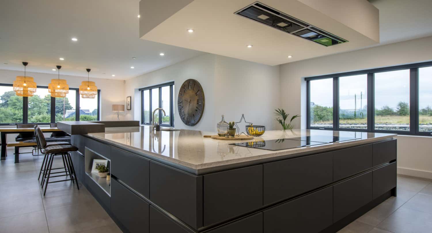 Modern kitchen with a large island, black cabinetry, pendant lights, and bar stools. Bespoke glass solutions, like expansive windows and glass roofing, flood the minimalist space with natural light and showcase lush greenery.