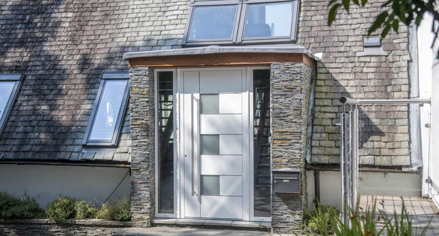 Modern white front door with geometric glass panels, framed by stone columns, on a house with grey shingles and large windows above the entrance. Shrubs line the pathway leading to the stylish front doors.