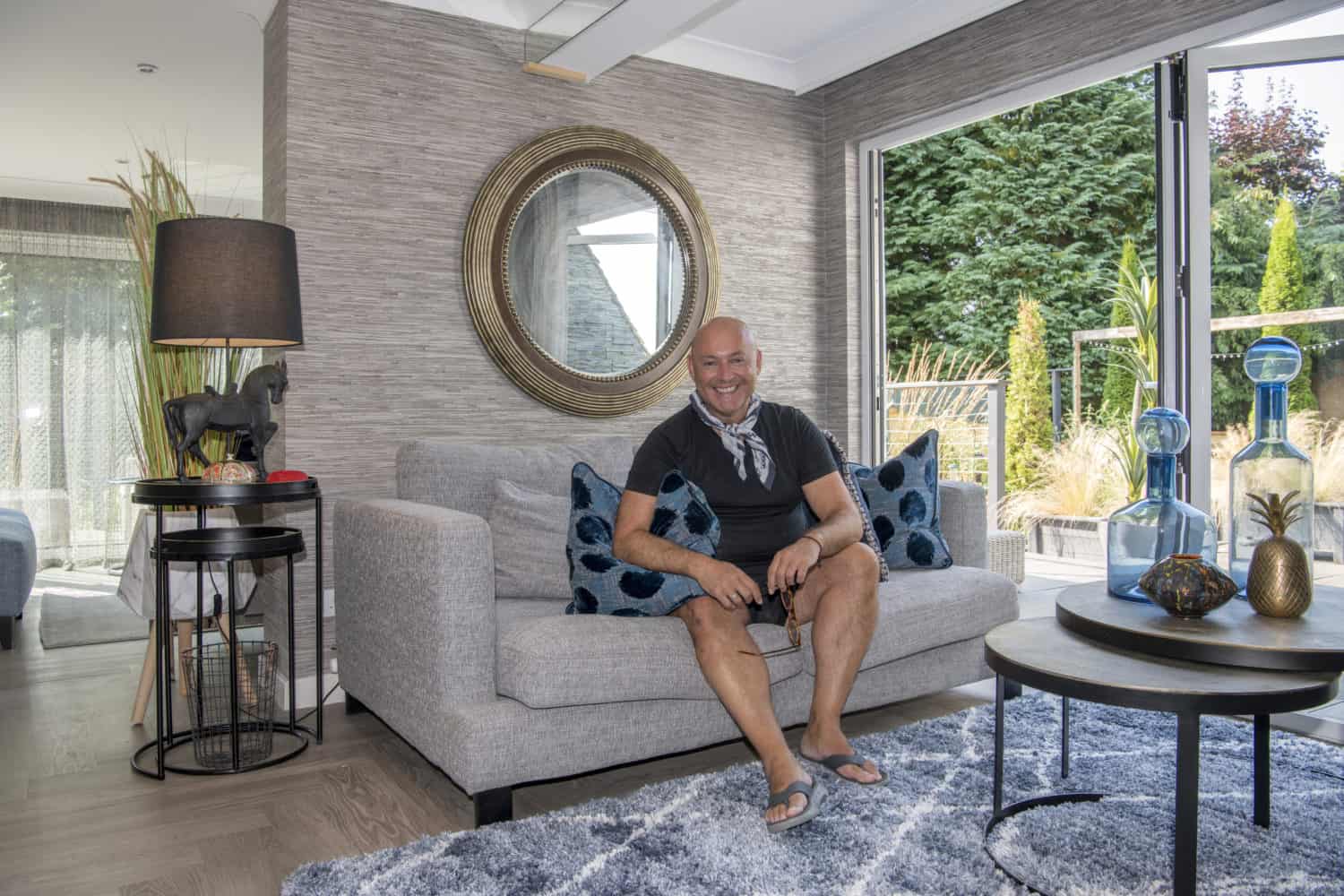 A man sits smiling on a light gray sofa with blue patterned pillows in a modern living room, decorated with glass vases, a round mirror, and large windows with sliding doors overlooking a green garden.