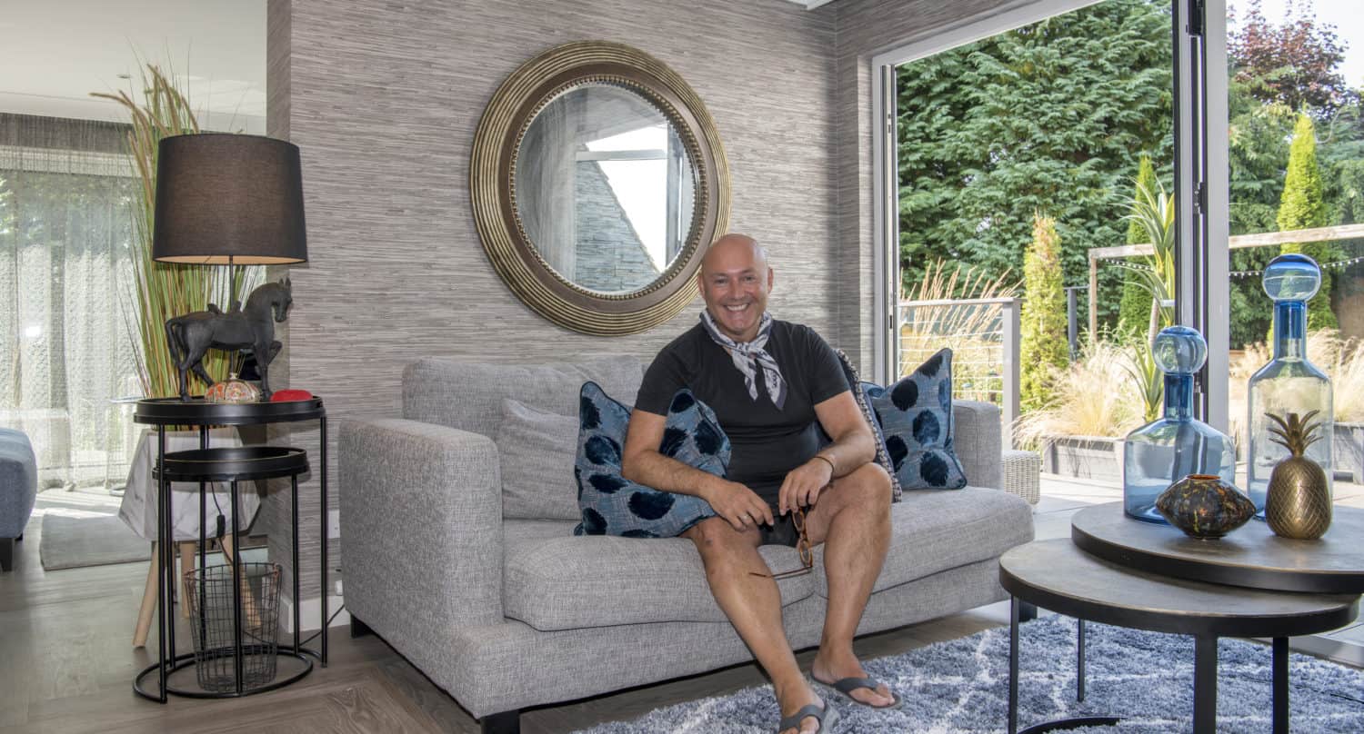 A man sits smiling on a light gray sofa with blue patterned pillows in a modern living room, decorated with glass vases, a round mirror, and large windows with sliding doors overlooking a green garden.
