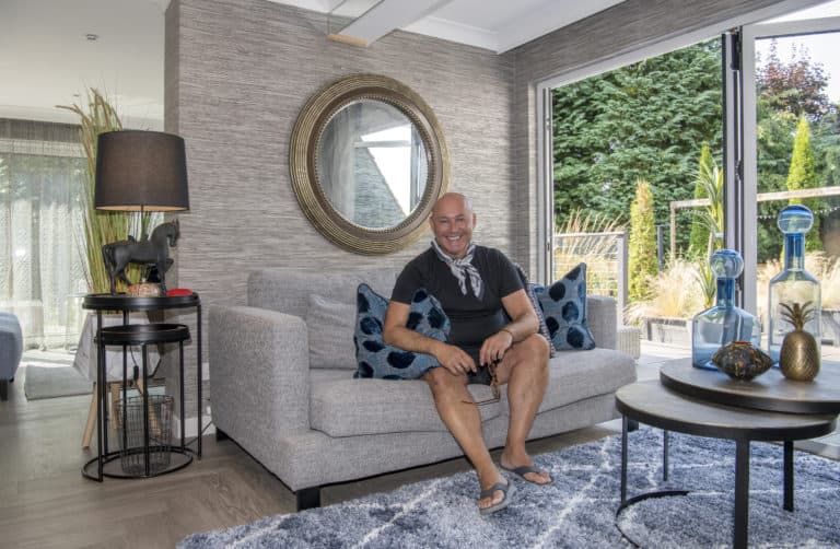 A man sits smiling on a light gray sofa with blue patterned pillows in a modern living room, decorated with glass vases, a round mirror, and large windows with sliding doors overlooking a green garden.