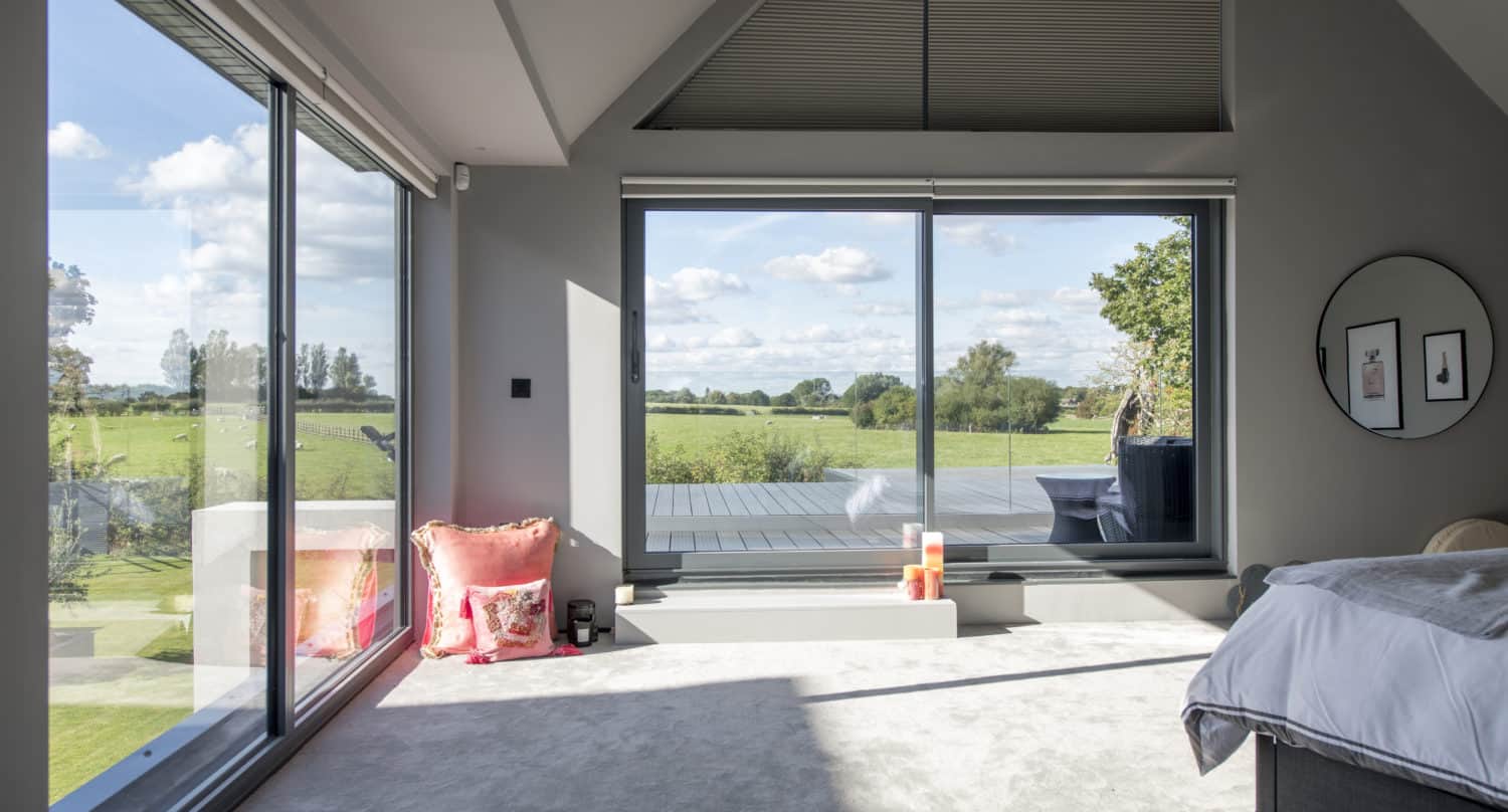 A modern bedroom with large floor-to-ceiling bespoke glass solutions overlooking a green field, letting in abundant natural light. The room features a bed, a round mirror, framed art, and decorative pillows and candles on the floor.