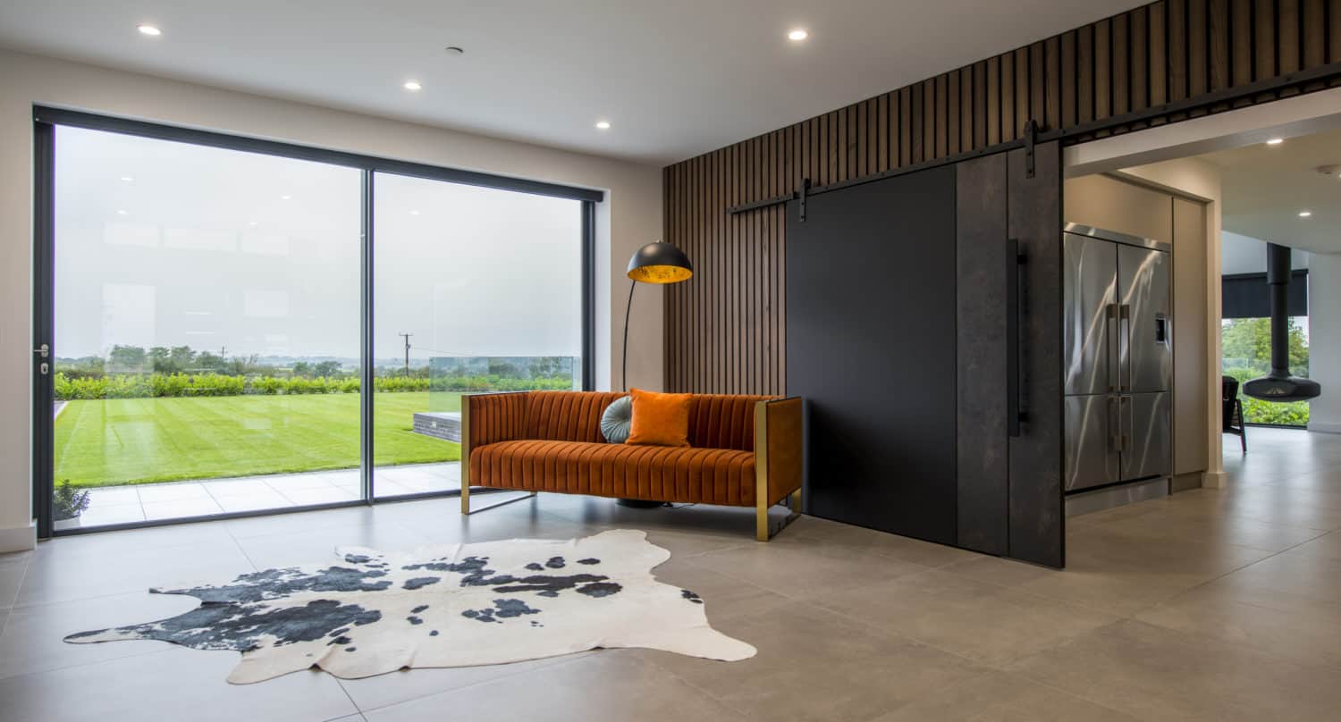Modern living space with large sliding glass doors, an orange patterned sofa, standing lamp, black bifold doors, and a cowhide rug on a tiled floor. The room overlooks a lush green lawn and features bespoke glass solutions throughout.