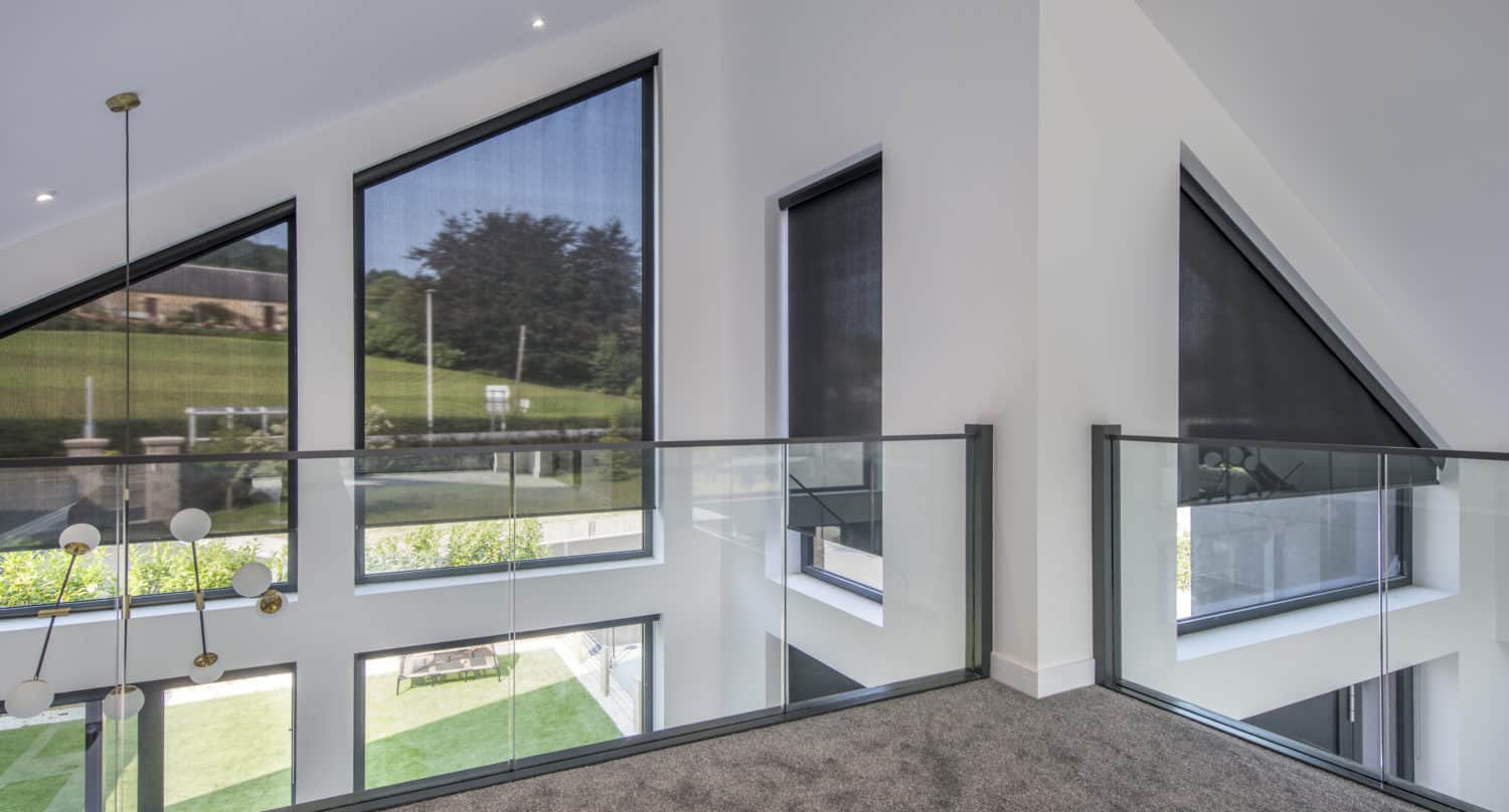 Modern interior with large triangular windows covered by dark blinds, glass railing, gray carpet, and a view of a green lawn and trees outside. Bespoke glass solutions enhance the bright space with white walls and contemporary lighting.