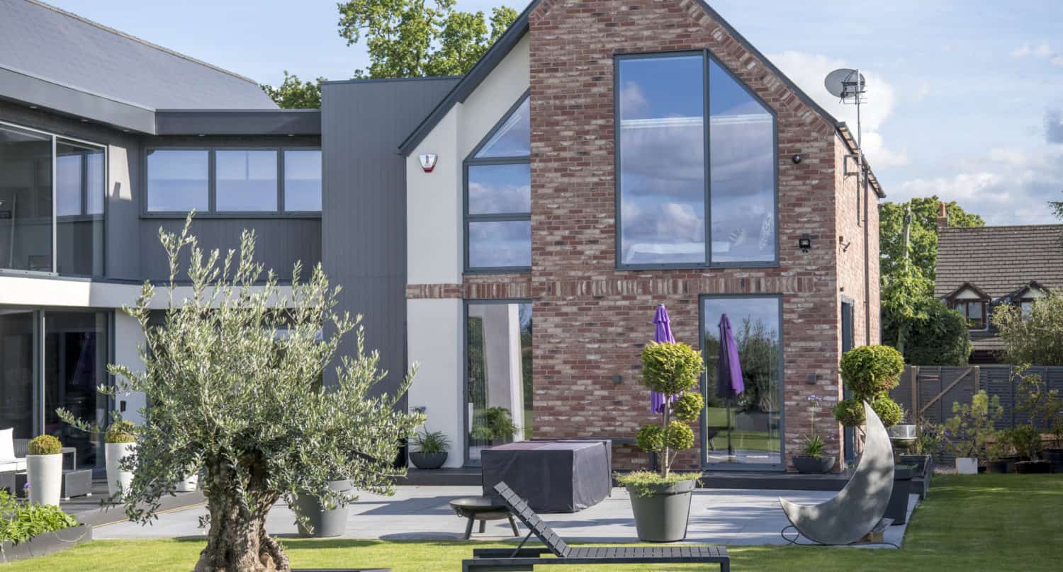 Modern house with large windows, brick accents, and sleek sliding doors, surrounded by a landscaped patio featuring lounge chairs, potted plants, and an olive tree. Purple umbrellas provide shade on a sunny day.