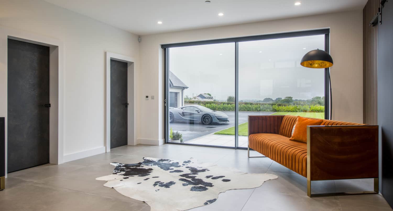 Modern living room with large floor-to-ceiling window, orange patterned sofa, black floor lamp, and a black-and-white cowhide rug. Sliding doors open to the outside, where a luxury car is parked with greenery and houses in the background.
