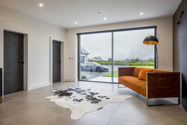 Modern living room with large floor-to-ceiling window, orange patterned sofa, black floor lamp, and a black-and-white cowhide rug. Sliding doors open to the outside, where a luxury car is parked with greenery and houses in the background.