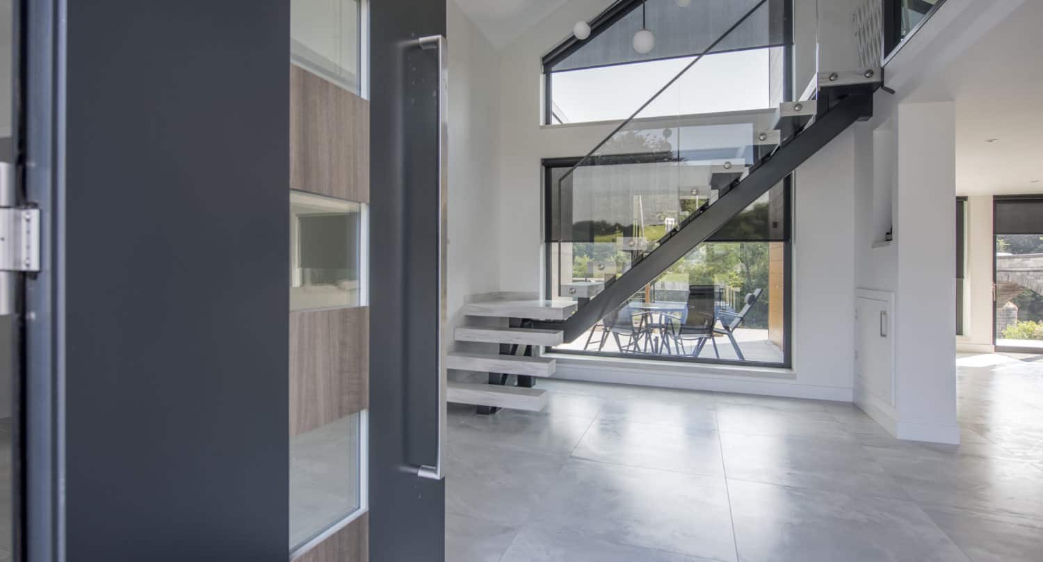 Modern interior with large tile flooring, a glass and metal staircase, tall windows flooding the space with natural light, and a glimpse of a dining area outside; bespoke glass solutions and a partially open front door are visible on the left.