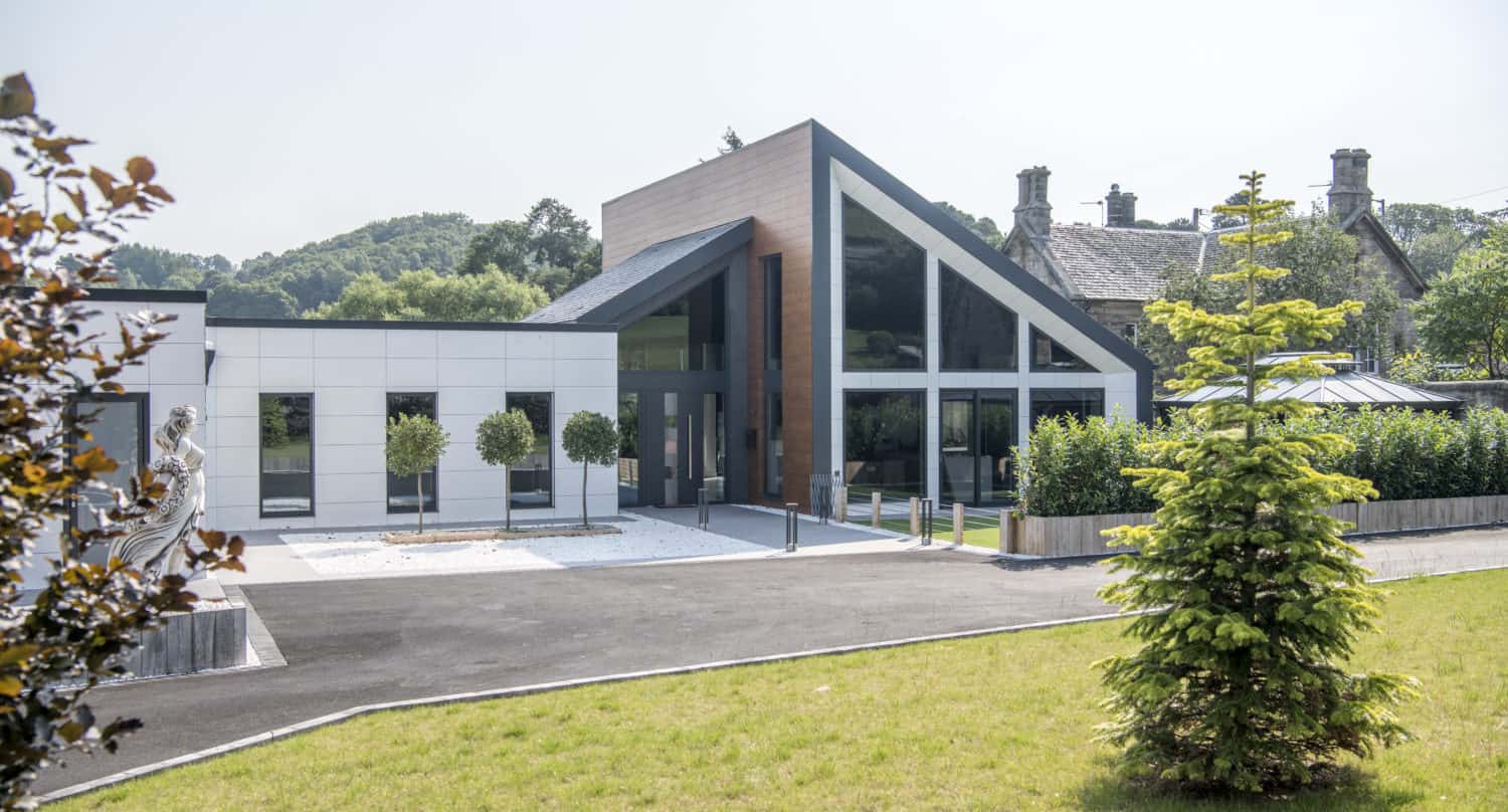 A modern building with large glass windows, sliding doors, and a slanted roof stands next to an older stone building, surrounded by green grass, trees, and landscaped shrubs on a sunny day.