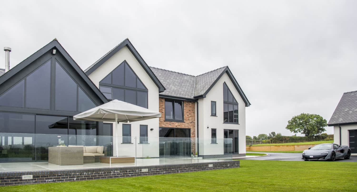 Modern two-story house with large glass windows, stylish sliding doors, a mix of white and brick exterior, and a spacious patio with outdoor seating and an umbrella. A sleek black car is parked on the driveway next to a well-kept lawn.