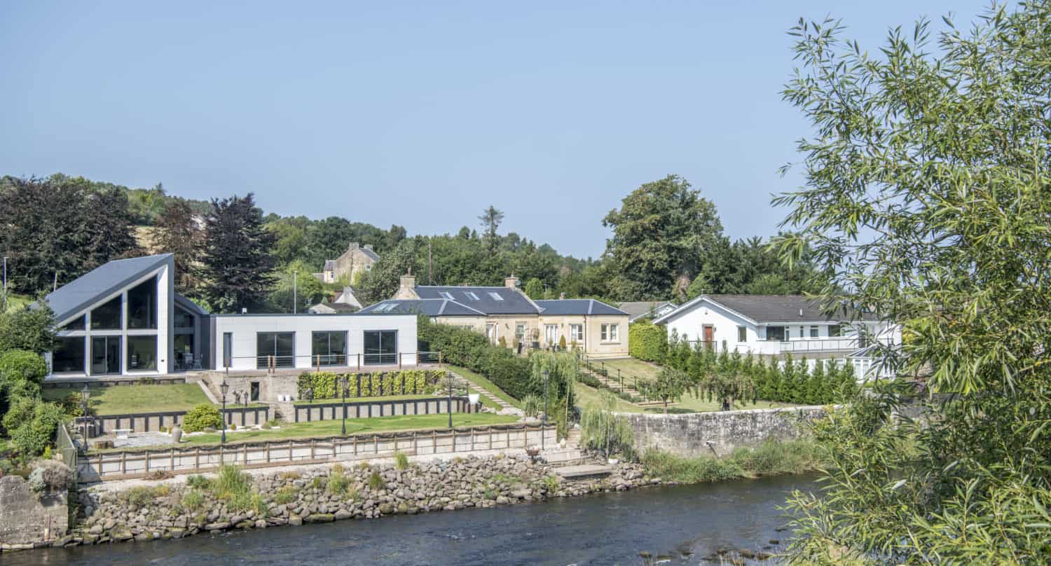 Modern and traditional houses with large windows, glass roofing, and manicured gardens stand on a grassy riverbank, bordered by trees and a stone wall, under a clear blue sky.