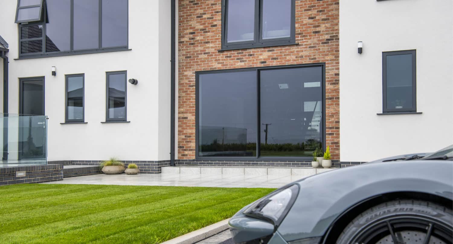 A modern house with large windows, bespoke glass solutions, a white and red brick exterior, neatly trimmed green lawn, and part of a sleek gray sports car parked in the driveway in the foreground.