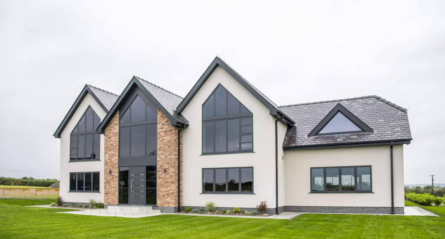 Modern two-story house with large windows, sliding doors, a brick and white exterior, gray roof, and well-kept green lawn under a cloudy sky.