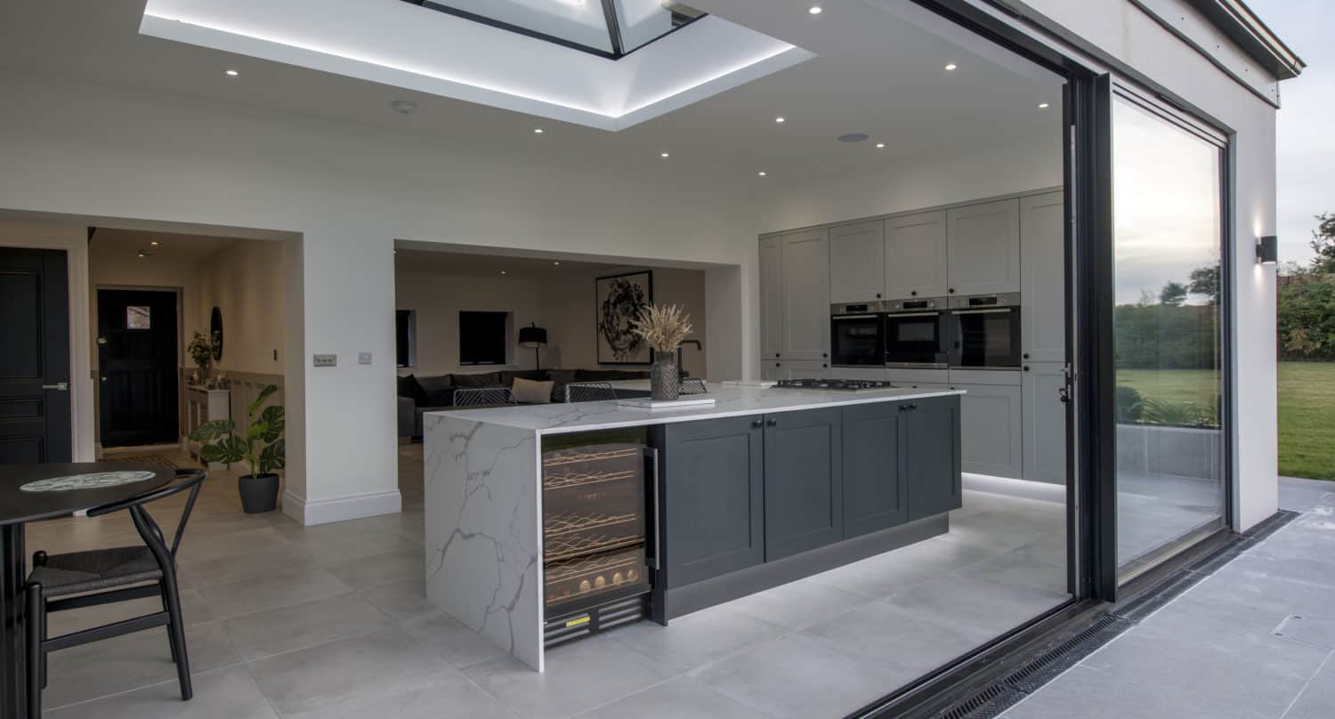 Modern open-plan kitchen with a large island, built-in wine fridge, grey cabinets, skylight, and bespoke glass sliding doors that open to a spacious green lawn. The space is sleek and well-lit with minimalist decor.