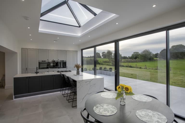Modern kitchen with glass roofing and large skylight, island with bar stools, built-in ovens, and floor-to-ceiling windows overlooking a green field. Round dining table with sunflowers in a vase is in the foreground.