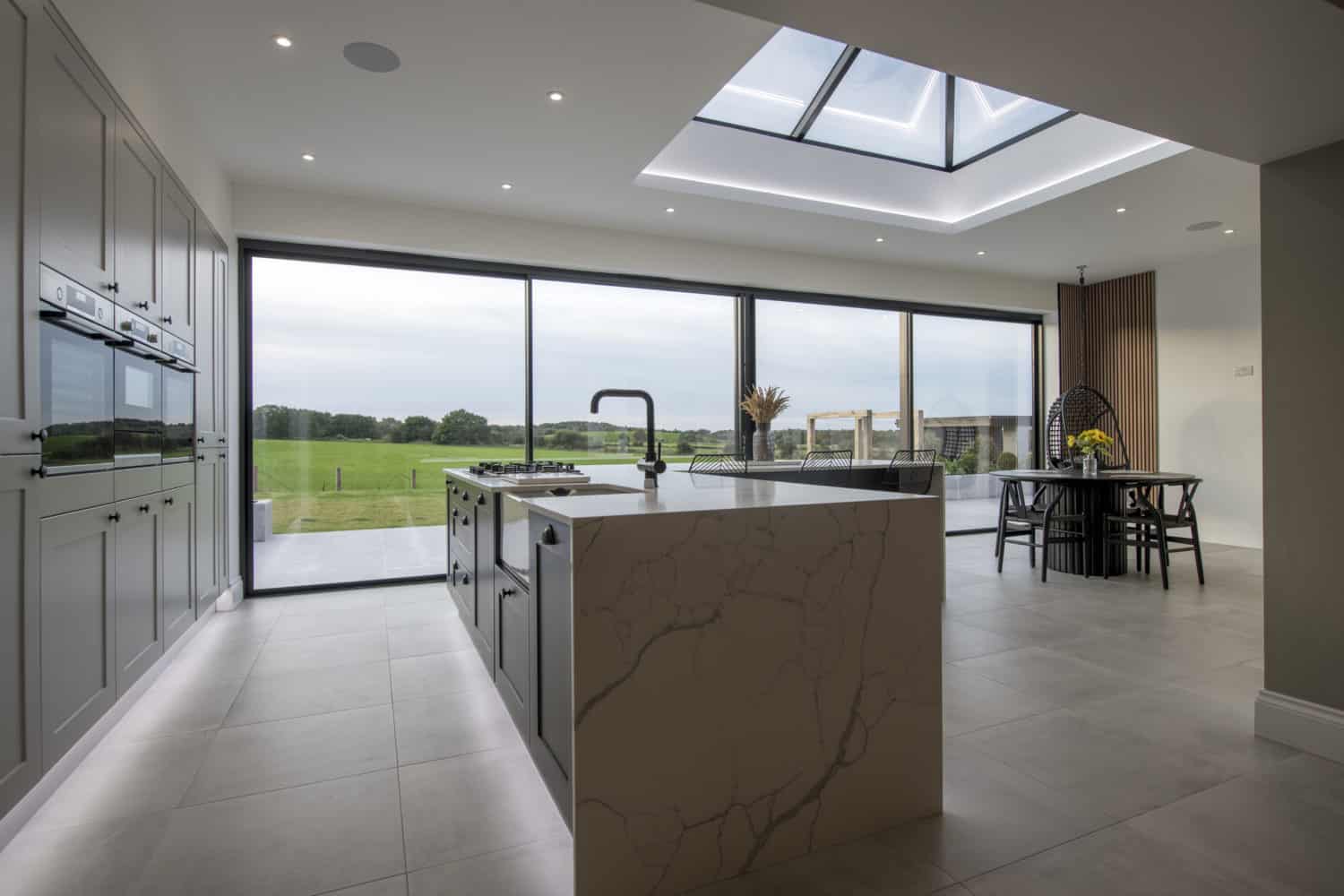 Modern kitchen with marble island, built-in appliances, large skylight, and bespoke glass solutions like floor-to-ceiling sliding doors opening to a scenic green landscape. Dining area with table and chairs is visible in the background.