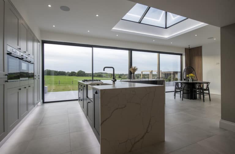 Modern kitchen with marble island, built-in appliances, large skylight, and bespoke glass solutions like floor-to-ceiling sliding doors opening to a scenic green landscape. Dining area with table and chairs is visible in the background.