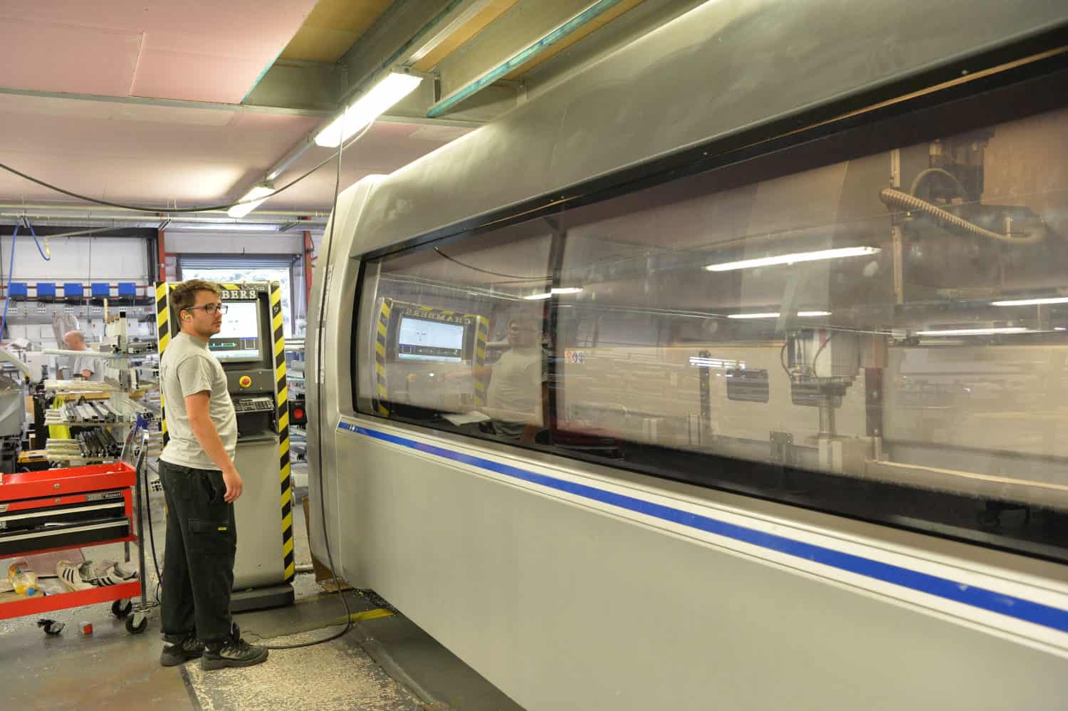 A man stands in a factory beside a large, gray industrial machine with a glass window and sliding doors, observing its operation. Shelves and tools are visible in the background.