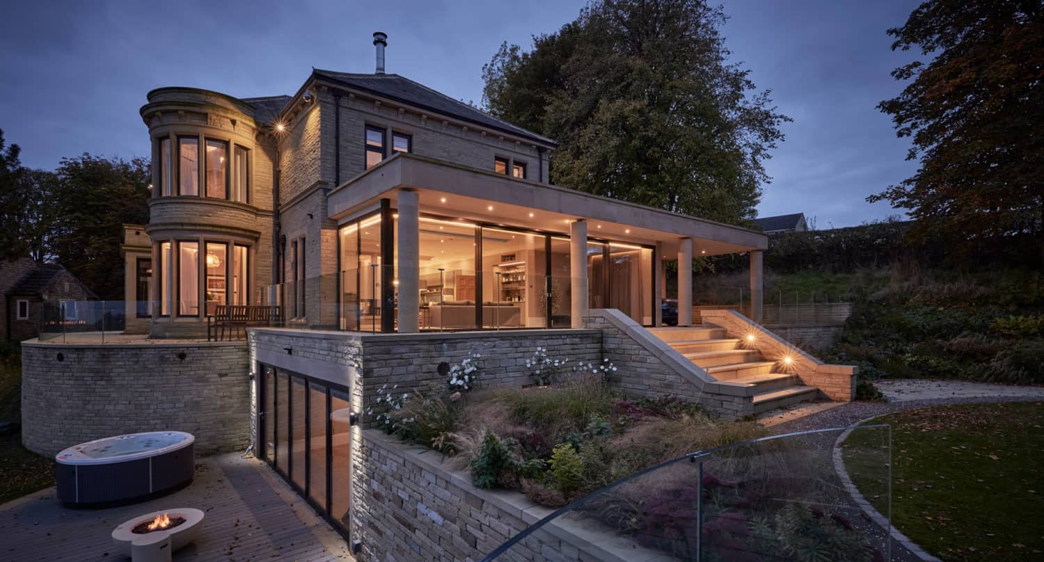 Modern, multi-level house with large glass windows and warm interior lighting at dusk, featuring sleek sliding doors, surrounded by landscaped gardens, stone walls, and an outdoor seating area with a round hot tub.