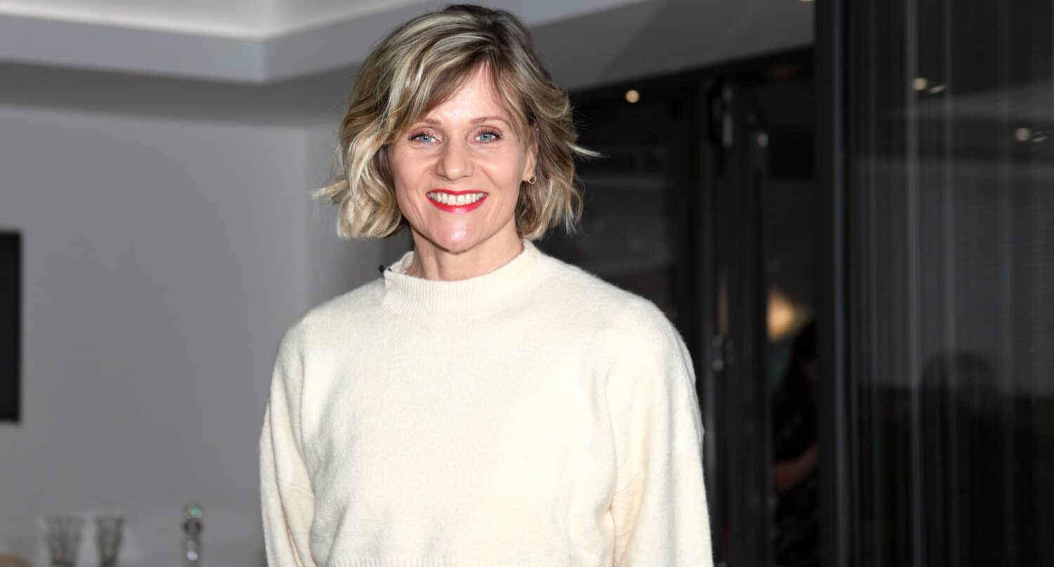 A woman with short blonde hair wearing a cream-colored sweater stands indoors, smiling at the camera. The background features modern decor and sleek sliding doors with dark glass.