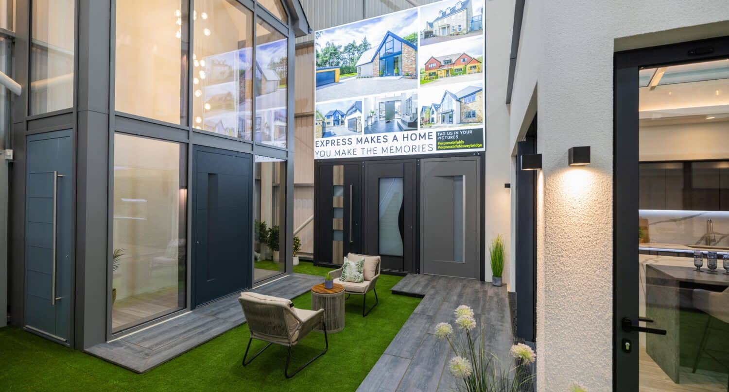 Modern indoor showroom with artificial grass, outdoor seating area, and potted plants. Large wall displays feature homes, while bifold doors and front doors enhance a bright, open space with bespoke glass solutions and high ceilings.