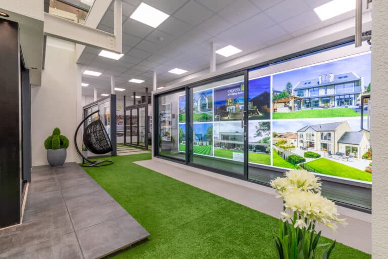 A modern indoor space with artificial grass flooring, potted plants, a black hanging chair, and a wall showcasing large photos of houses—enhanced by bespoke glass solutions and bright ceiling lights.