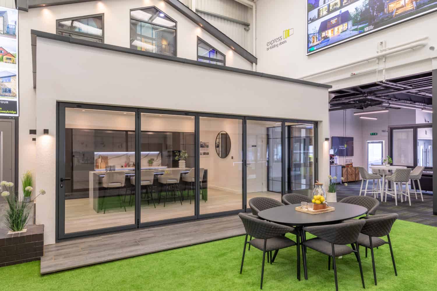 A modern showroom with large bifold doors, an indoor patio area featuring a round table and chairs on artificial grass, and a stylish meeting room visible inside. Bright, open atmosphere enhanced by bespoke glass solutions and decorative plants.