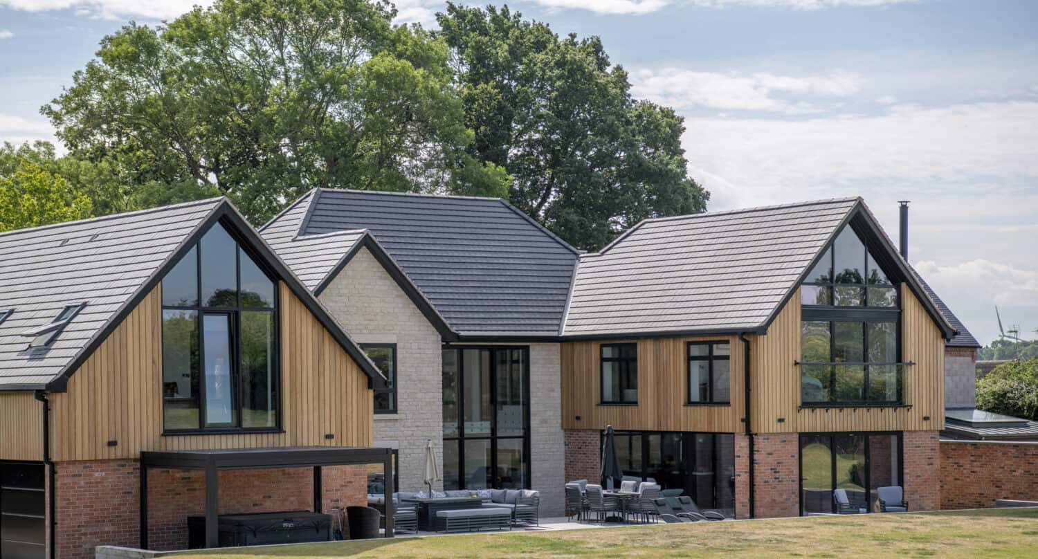 Modern two-story house with large windows, bifold doors, wooden and brick exterior, gabled roofs, and a spacious outdoor patio, surrounded by grass and trees under a partly cloudy sky.