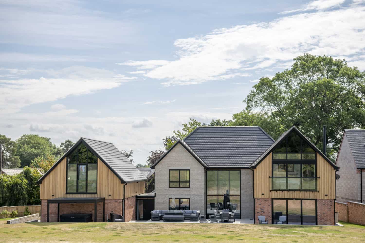 A modern two-story house with bespoke glass solutions, large front doors, light wood and brick exterior, and multiple steeply pitched roof sections, set on a grassy lawn with trees in the background under a partly cloudy sky.