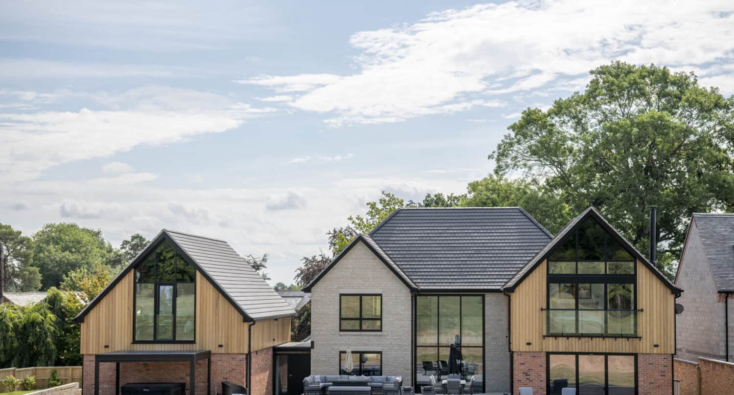 A modern two-story house with bespoke glass solutions, large front doors, light wood and brick exterior, and multiple steeply pitched roof sections, set on a grassy lawn with trees in the background under a partly cloudy sky.