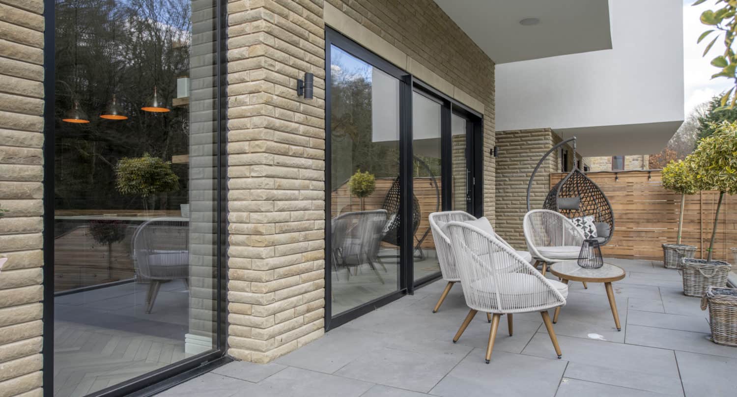 Modern patio with light gray tile flooring, wicker chairs around a round wooden table, a hanging chair, potted plants, and large bespoke glass solutions or sliding doors reflecting trees and interior lights.