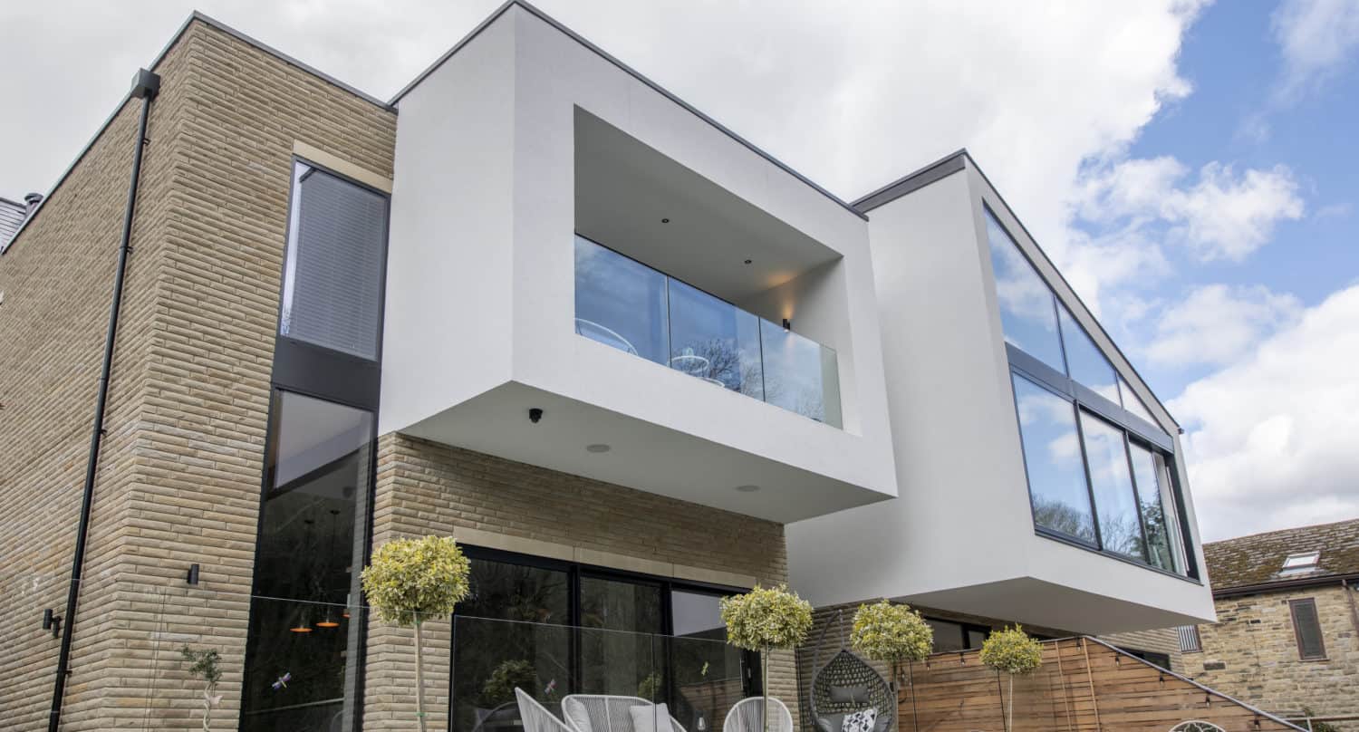 Modern two-story house with brick and white geometric exterior, large glass windows, bespoke glass solutions for railings, inviting front doors, balconies, patio seating, potted topiary trees—all under a partly cloudy sky.