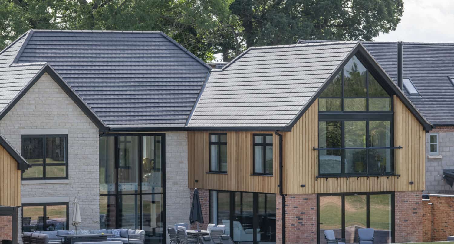 Modern two-story house with a mix of brick, wood, and stone exterior, floor-to-ceiling windows, bespoke glass solutions like glass roofing, an outdoor seating area, and surrounded by green trees and grass.
