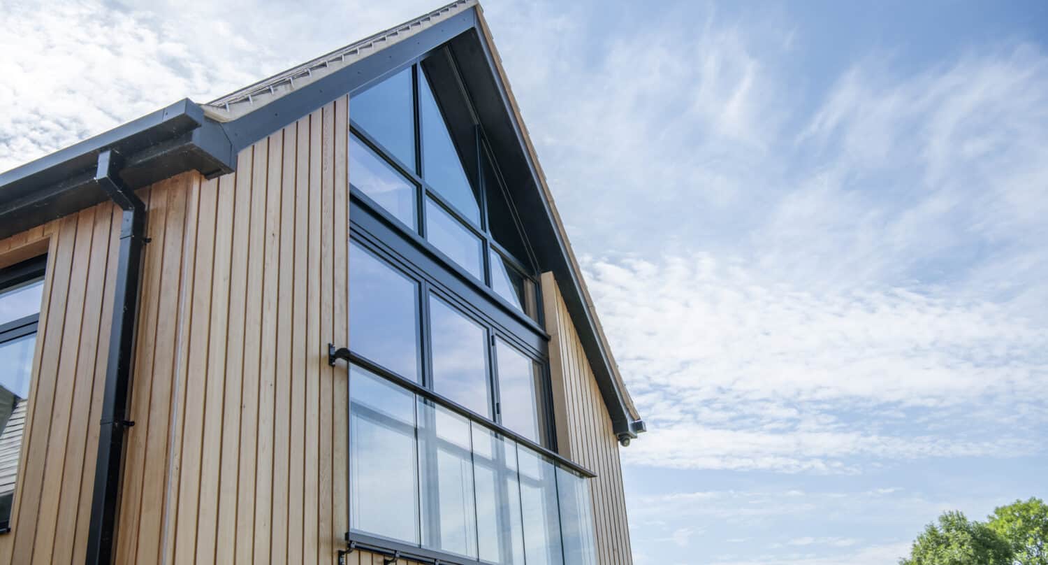 Modern house with vertical wooden siding and large glass windows, featuring bespoke glass solutions and sliding doors, viewed from a low angle against a bright blue sky with scattered clouds; a tree is partially visible on the right.