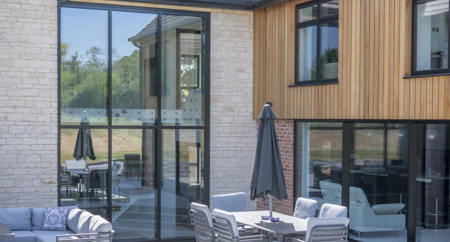 Modern house exterior with large floor-to-ceiling windows, wood and stone walls, and a patio area featuring sliding doors, a gray outdoor sofa, dining table with chairs, and closed umbrella. Trees are visible in the background.