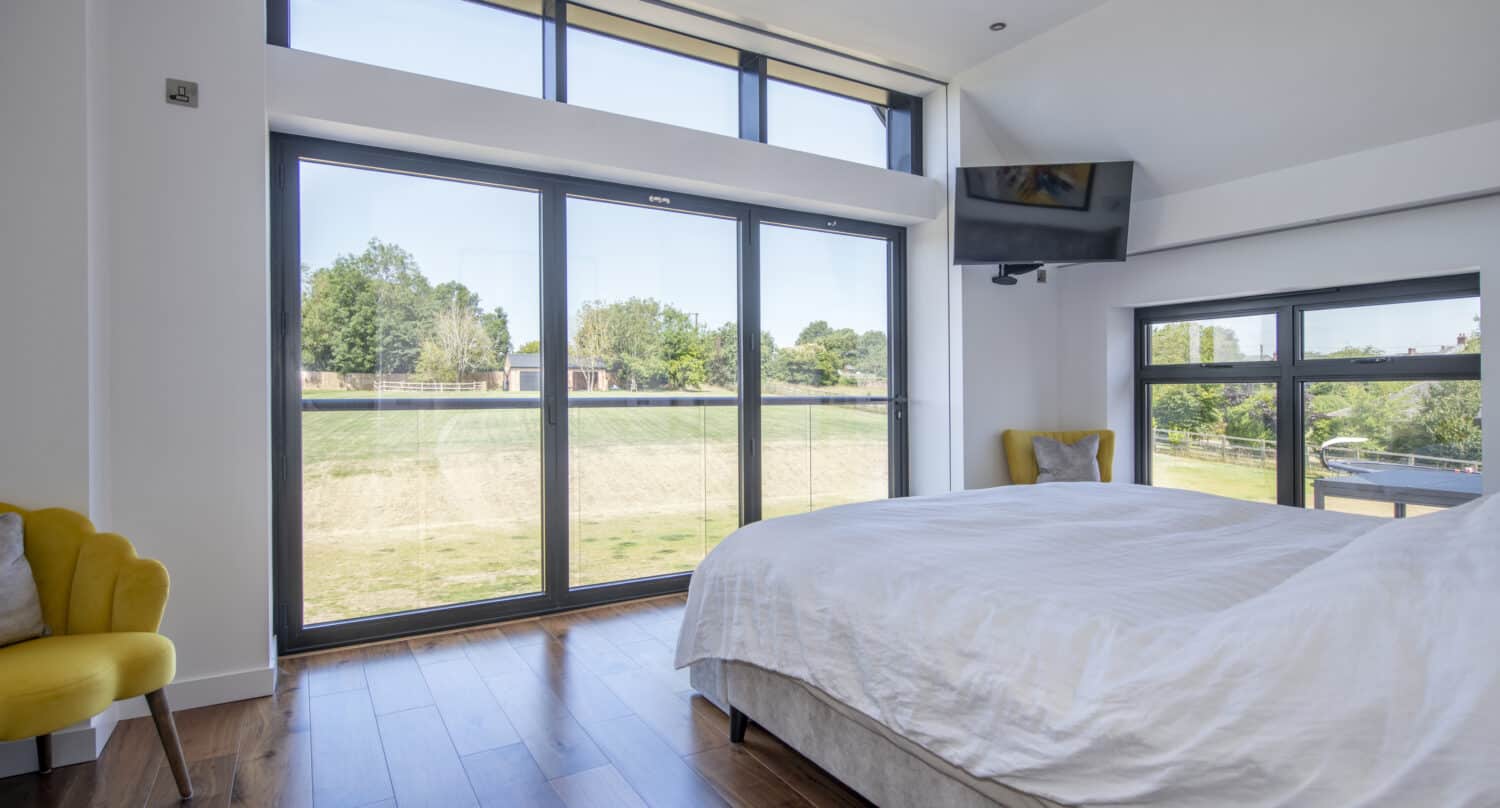 Modern bedroom with large floor-to-ceiling bespoke glass solutions, a neatly made bed with white bedding, wooden floors, two yellow armchairs, and a wall-mounted TV; green lawn and trees visible outside.