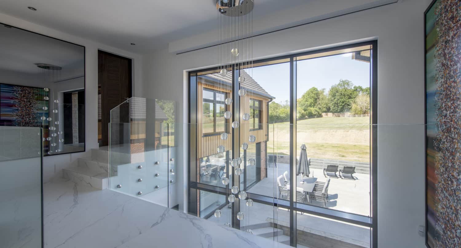 Modern interior with large windows and sliding doors overlooking a patio with outdoor seating. White marble floors, glass railing, and a contemporary hanging light fixture enhance the bright, open space. Trees are visible outside.