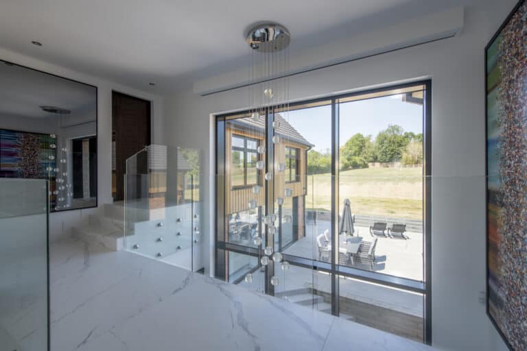 Modern interior with large windows and sliding doors overlooking a patio with outdoor seating. White marble floors, glass railing, and a contemporary hanging light fixture enhance the bright, open space. Trees are visible outside.