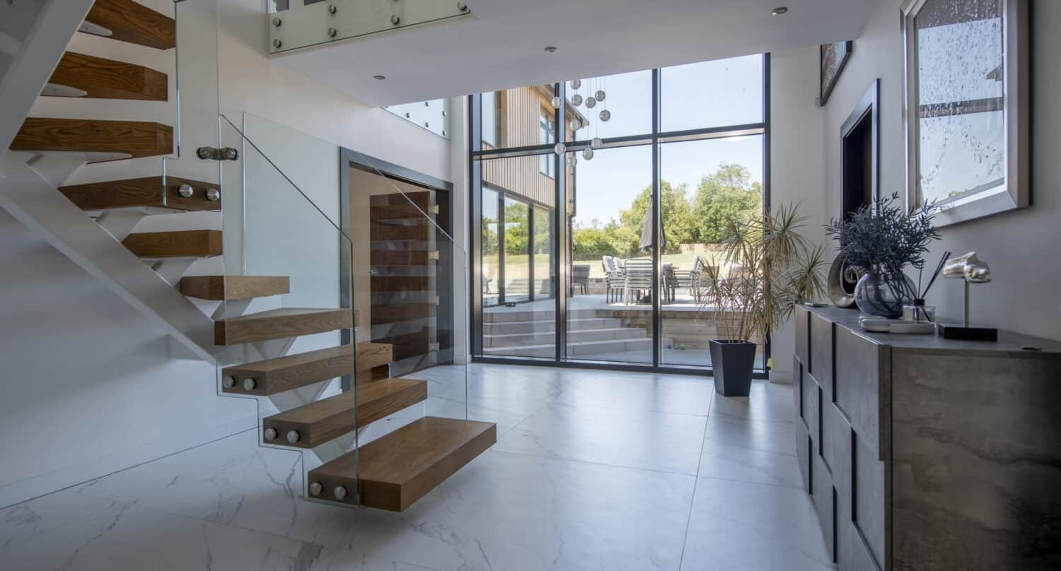 Modern entryway with floating wooden staircase, glass railings, large floor-to-ceiling windows, white marble tile floor, and a console table with decorative items; sliding doors open to an outdoor patio visible through the windows.