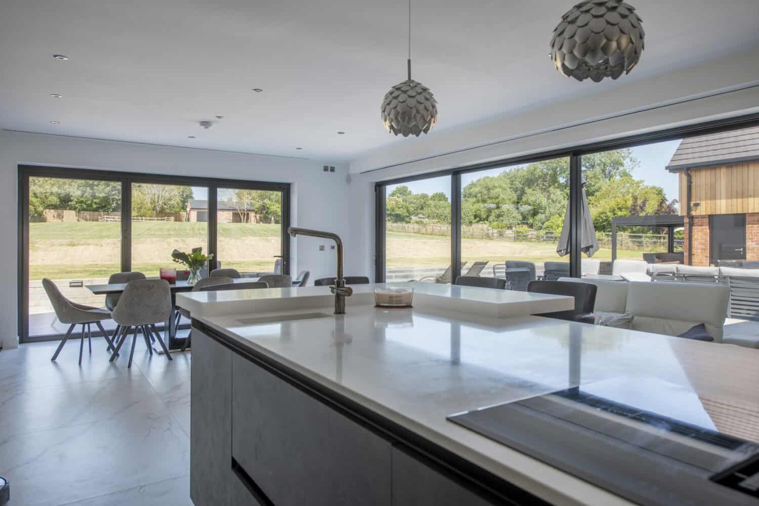 Modern open-plan kitchen and dining area with large island, pendant lights, and floor-to-ceiling bespoke glass solutions, including sliding doors overlooking a patio and grassy outdoor space.