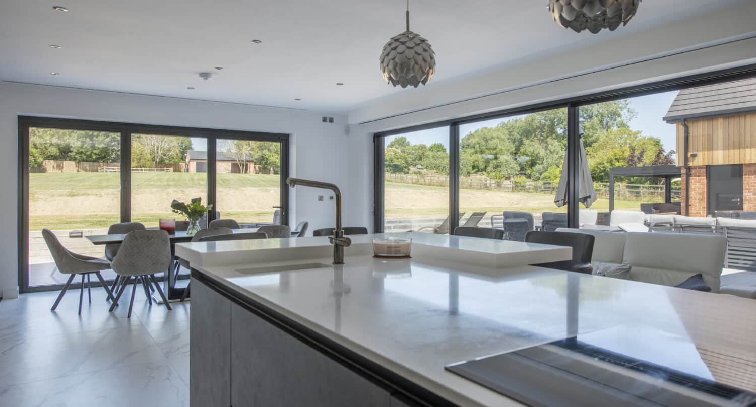 Modern open-plan kitchen and dining area with large island, pendant lights, and floor-to-ceiling bespoke glass solutions, including sliding doors overlooking a patio and grassy outdoor space.