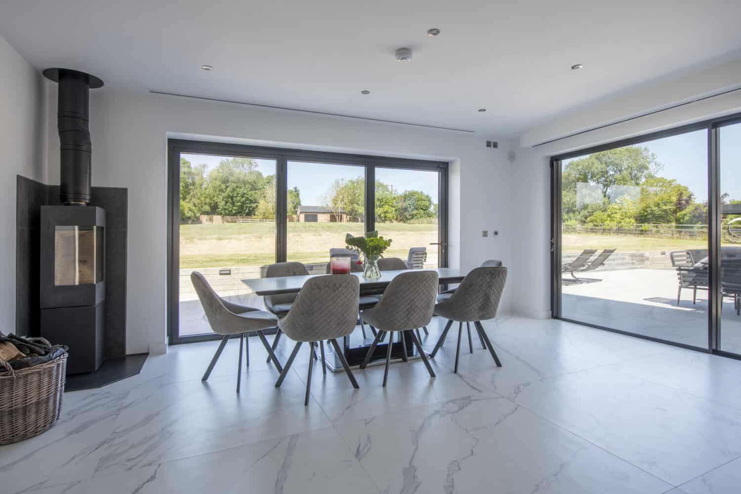 Modern dining area with a bespoke glass table and six gray chairs on white marble flooring. Large sliding glass doors open to a sunny patio with outdoor seating and green lawns. A black wood stove sits in the corner.