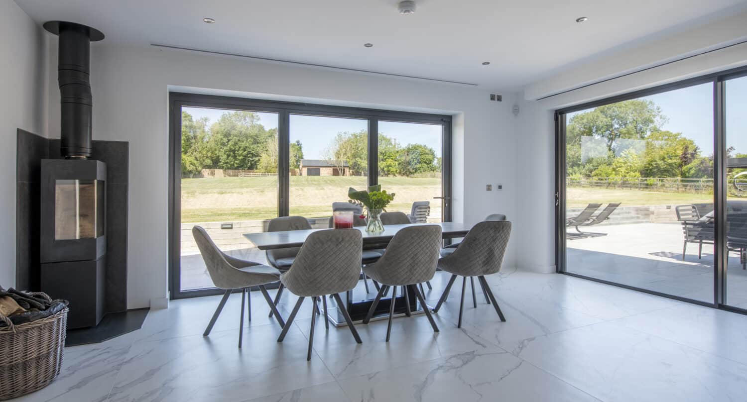 Modern dining area with a bespoke glass table and six gray chairs on white marble flooring. Large sliding glass doors open to a sunny patio with outdoor seating and green lawns. A black wood stove sits in the corner.