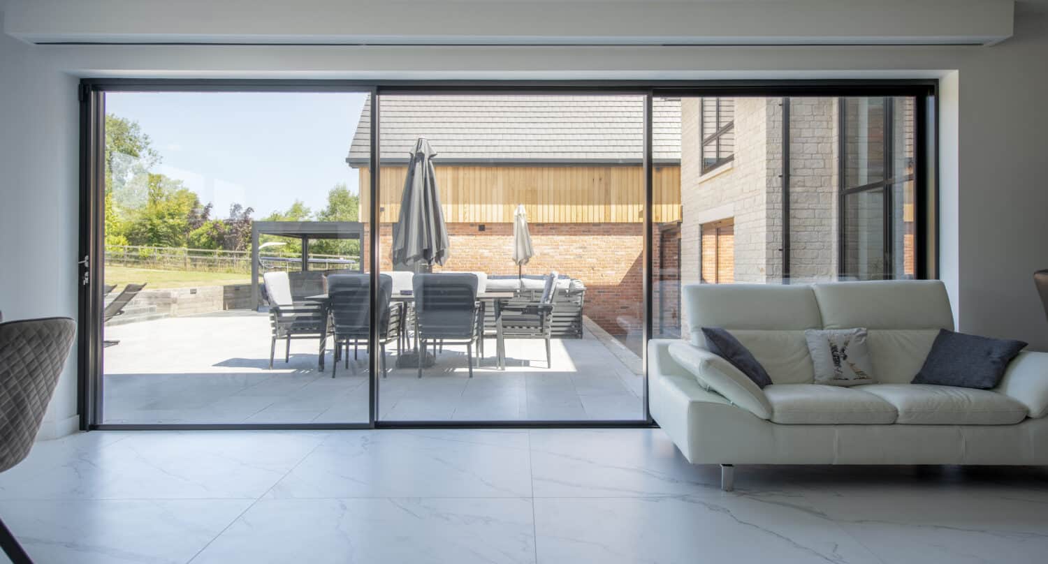 A modern living room with a white sofa and large bifold doors that open to a patio with a dining table, chairs, and closed umbrellas. The outdoor area is surrounded by trees and a contemporary house.