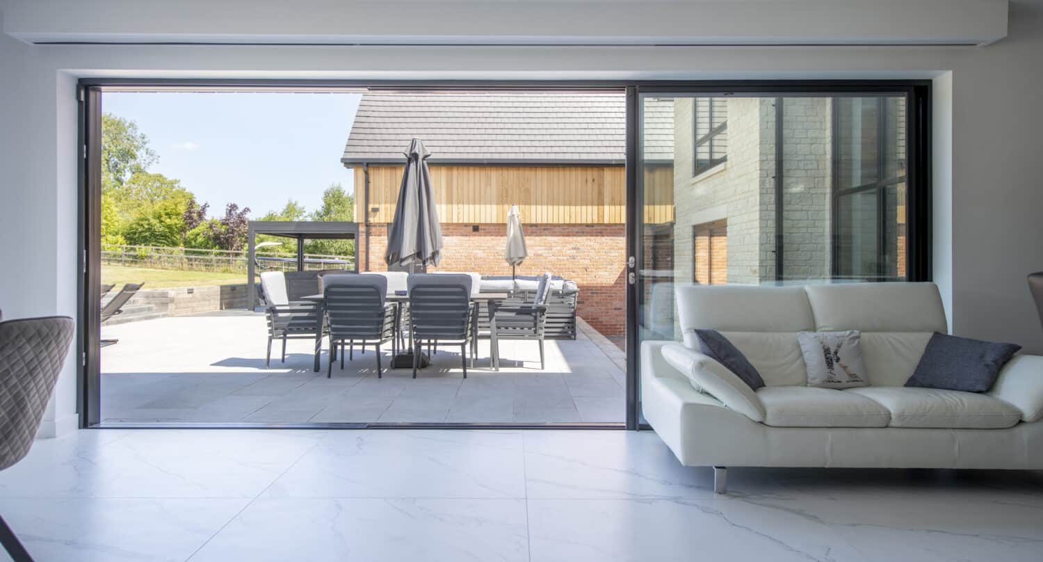 Modern living room with a white sofa and stylish bifold doors opening to a patio with outdoor dining furniture, umbrellas, and a view of a wooden building and greenery outside.