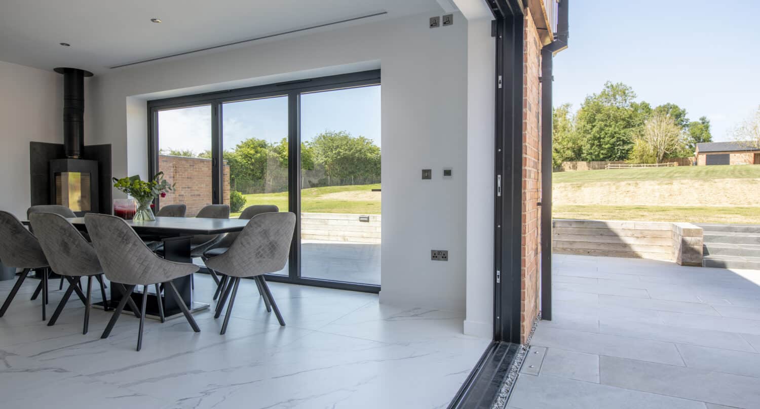 Modern dining area with a black table and gray chairs, large sliding doors open to a spacious patio and grassy backyard with trees under a clear sky. Marble floors inside transition to tiled flooring outside, enhanced by bespoke glass solutions.