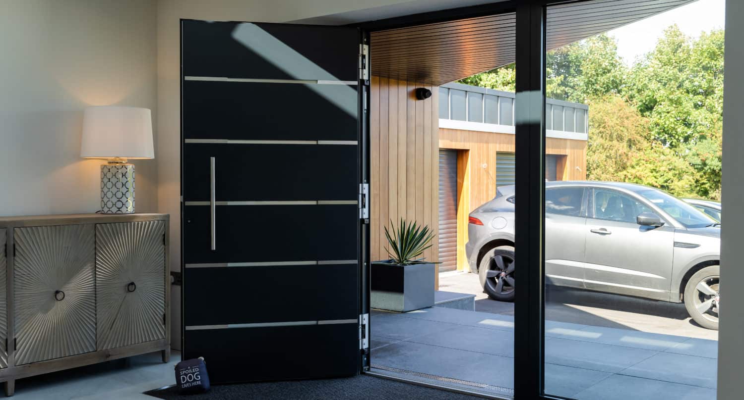 A modern front door is open, revealing a driveway with a gray car parked outside. Inside, there’s a stylish cabinet with a lamp and bifold doors nearby. Sunlight streams in through large windows onto a doormat with a sign that reads DOG.